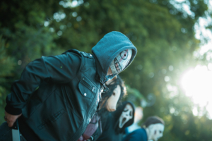 Ein Teilnehmer des Elbenwald Festivals trägt eine auffällige Maske und eine schwarze Lederjacke. Im Hintergrund sind weitere verkleidete Personen zu sehen, die die kreative und fantasievolle Atmosphäre des Festivals widerspiegeln.