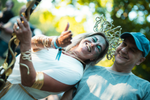 Zwei fröhliche Festivalbesucher beim Elbenwald Festival, gekleidet in fantasievolle Kostüme. Die Frau trägt eine auffällige Kopfbedeckung und hat glitzerndes Make-up, während der Mann lächelt und entspannt wirkt. Im Hintergrund sind weitere Teilnehmer in festlicher Stimmung zu sehen.