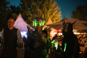 Zwei verkleidete Teilnehmer des Elbenwald Festivals stehen in der Dämmerung. Der erste trägt eine traditionelle Tracht, während der zweite in einer beeindruckenden, leuchtenden Rüstung mit spitzen Hörnern zu sehen ist. Im Hintergrund sind festliche Zelte und Besucher zu erkennen.