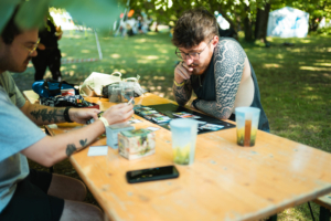 Zwei Männer sitzen an einem Holztisch beim Elbenwald Festival und spielen ein Kartenspiel. Auf dem Tisch liegen verschiedene Spielkarten, Getränke und persönliche Gegenstände. Im Hintergrund sind grüne Bäume und Festivalzelte sichtbar.