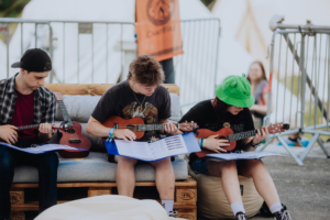 Ukulele Drei junge Menschen spielen Ukulele auf einem Sofa beim Elbenwald Festival. Sie sitzen mit Notenblättern und konzentrieren sich auf ihre Musik. Im Hintergrund sind Zelte und eine Absperrung zu sehen, was die Festivalatmosphäre unterstreicht.