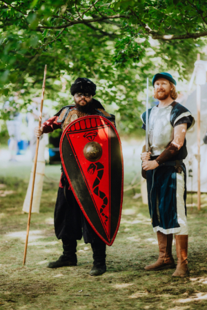 Zwei verkleidete Teilnehmer des Elbenwald Festivals stehen im Freien. Der erste trägt ein rotes Schild und einen Speer, während der zweite in einer Rüstung mit Schwert posiert. Die Umgebung ist von Bäumen umgeben, was eine festliche Atmosphäre schafft.