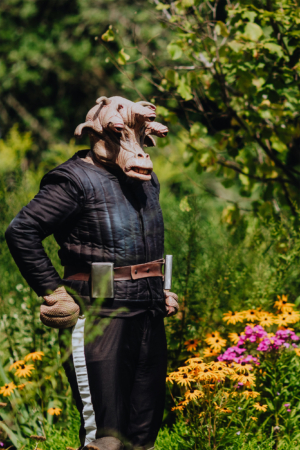 Ein Teilnehmer des Elbenwald Festivals in einem kreativen Kostüm mit einem Tierkopf, umgeben von bunten Blumen und üppigem Grün, das die lebendige Atmosphäre des Festivals widerspiegelt. Ideal für Fans von Fantasy und Science-Fiction.