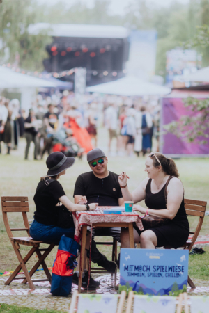 Drei Freunde sitzen an einem Tisch und spielen Spiele auf dem Elbenwald Festival. Im Hintergrund sind Menschenmengen und die Bühne zu sehen. Ein Schild mit der Aufschrift „Mitmach Spielwiese: Tummeln, Spielen, Chillen“ steht auf dem Tisch.