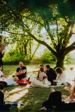 Gruppengespräch unter einem Baum beim Elbenwald Festival, wo Teilnehmer in kreativen Kostümen zusammenkommen. Die entspannte Atmosphäre und das grüne Umfeld fördern den Austausch von Ideen und Begeisterung für Fantasy und Musik.