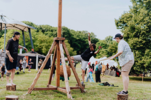 Drei Festivalbesucher beim Elbenwald Festival versuchen, ihr Gleichgewicht auf einem Holzstamm zu halten. Im Hintergrund sind andere Teilnehmer und Zelte zu sehen, die die lebendige Atmosphäre des Events unterstreichen.