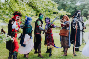 Bunte Cosplay-Gruppe auf dem Elbenwald Festival, bestehend aus Charakteren mit kreativen Kostümen, darunter Pirat, Elfe und Krieger, die in einer lebhaften Umgebung stehen, umgeben von Bäumen und einem Teich im Hintergrund.