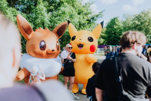 Zwei große, aufblasbare Pokémon-Figuren, Pikachu und Evoli, stehen auf dem Elbenwald Festival. Eine Frau mit Sonnenbrille hält ein Mikrofon und interagiert mit den Festivalbesuchern, während im Hintergrund andere Teilnehmer zu sehen sind.