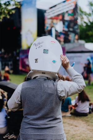 Eine Person mit einem Toilettensitz auf dem Kopf, dekoriert mit Aufklebern, steht auf dem Elbenwald Festival. Im Hintergrund sind Festivalbesucher und eine Bühne zu sehen, die eine lebendige Atmosphäre des Events widerspiegelt.