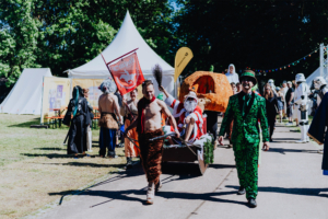 Bunte Parade beim Elbenwald Festival mit kostümierten Teilnehmern, darunter ein Mann im grünen Anzug, eine Person im Weihnachtsmann-Kostüm und weitere Charaktere aus der Fantasiewelt. Im Hintergrund sind Festzelte und Zuschauer zu sehen.