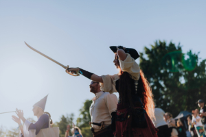 Eine Teilnehmerin des Elbenwald Festivals in Piratenkostüm hält ein Schwert in der Hand. Im Hintergrund sind weitere Festivalbesucher zu sehen, die in kreativen Kostümen applaudieren. Die Szene spielt sich bei strahlendem Sonnenschein ab.