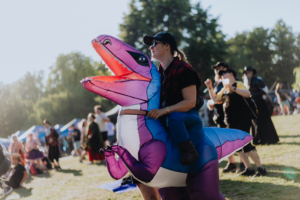 Eine Person in einem aufblasbaren Dinosaurier-Kostüm reitet auf dem Elbenwald Festival, umgeben von fröhlichen Festivalbesuchern. Die Szene zeigt die lebendige Atmosphäre der Veranstaltung mit Musik, Cosplay und kreativen Kostümen.