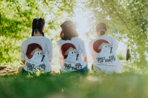 Drei Personen sitzen im Gras mit dem Rücken zur Kamera, tragen weiße T-Shirts mit einem grafischen Design und dem Schriftzug "Fight for the Party". Die Szene ist von Bäumen umgeben und vermittelt eine entspannte Festivalatmosphäre beim Elbenwald Festival.