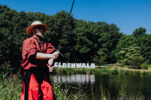 Ein Teilnehmer in historischer Kleidung mit einem Schwert posiert am Ufer eines Sees beim Elbenwald Festival, im Hintergrund steht das große, weiße Festival-Logo vor einer grünen Landschaft unter strahlend blauem Himmel.