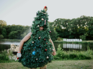 Ein Festivalbesucher verkleidet sich als Weihnachtsbaum mit bunten Ornamenten, während er am Ufer eines Sees steht. Im Hintergrund ist das Elbenwald-Logo sichtbar, das die fröhliche und kreative Atmosphäre des Festivals unterstreicht.