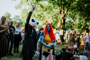 Zwei Cosplayer, die als Jack Skellington und Sally aus "Die Nightmare Before Christmas" verkleidet sind, posieren fröhlich auf dem Elbenwald Festival. Im Hintergrund sind weitere Festivalbesucher inmitten einer grünen, lebhaften Umgebung zu sehen.