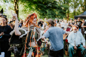Menschen auf dem Elbenwald Festival tanzen und feiern in farbenfrohen Kostümen. Eine Frau mit roten Haaren und Blumenkranz lächelt fröhlich, während andere Besucher im Hintergrund applaudieren und die festliche Atmosphäre genießen.