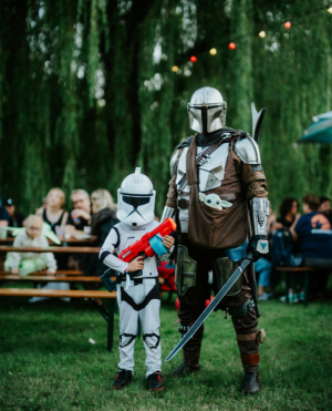 Zwei Cosplayer in beeindruckenden Star Wars-Outfits auf dem Elbenwald Festival. Ein Kind im Stormtrooper-Kostüm hält eine Spielzeugwaffe, während der Erwachsene als Mandalorianer verkleidet ist. Bunte Lichter und Festivalbesucher im Hintergrund verleihen eine lebendige Atmosphäre.