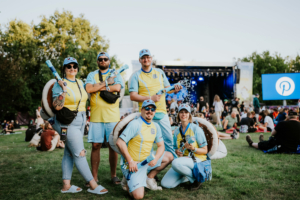 Fünf Teilnehmer in bunten Kostümen mit Wasserpistolen und Accessoires stehen auf dem Elbenwald Festival. Im Hintergrund sind die Bühne und zahlreiche Besucher zu sehen. Die fröhliche Atmosphäre verdeutlicht das kreative und gemeinschaftliche Erlebnis des Festivals.