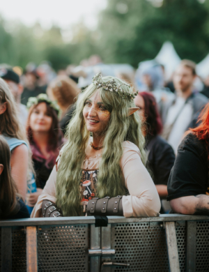 Eine lächelnde Frau mit langen, grünen Haaren und Elfenohren, geschmückt mit einer Blumenkrone, steht an einem Absperrgitter beim Elbenwald Festival. Im Hintergrund sind weitere Festivalbesucher in bunten Outfits zu sehen, die die Atmosphäre genießen.