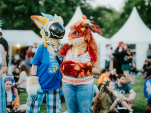 Zwei Personen in kreativen Tierkostümen stehen auf dem Elbenwald Festival, umgeben von Zeltständen und einer fröhlichen Menschenmenge. Die Atmosphäre ist lebhaft und bunt, geprägt von Musik und Fantasie.