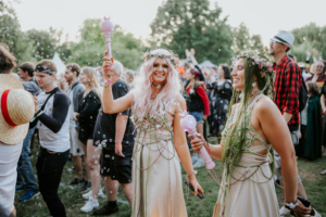 Zwei Frauen in fantasievollen Kostümen mit Blumenkränzen und glitzernden Accessoires feiern auf dem Elbenwald Festival. Sie halten bunte Zauberstäbe und umgeben von fröhlichen Festivalbesuchern, während Seifenblasen in der Luft schweben.