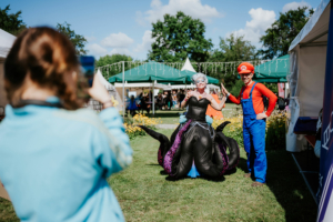 Kostümierte Besucher auf dem Elbenwald Festival posieren für ein Foto. Eine Frau in einem kreativen Ursula-Kostüm und ein Mann als Mario stehen im Vordergrund, während im Hintergrund Festivalatmosphäre mit Zelten und Blumen herrscht.