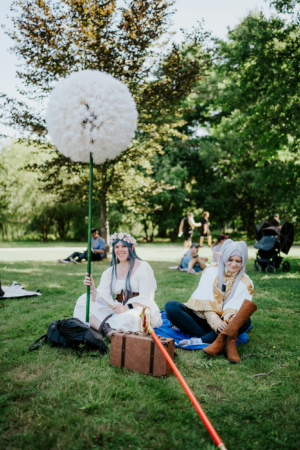 Zwei Cosplayerinnen im Grünen beim Elbenwald Festival, eine mit einem großen, weißen Pusteblumen-Stab und Blumenkranz, die andere in einem goldenen Outfit. Im Hintergrund sind weitere Festivalbesucher und Bäume zu sehen.