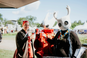 Drei Festivalbesucher in Kostümen halten bunte Getränke in der Hand. Die Frau in traditioneller Kleidung hat rote Haare, während die beiden anderen in weißen Masken und bunten Accessoires gekleidet sind. Im Hintergrund sind Festivalzelte und ein blauer Himmel zu sehen.