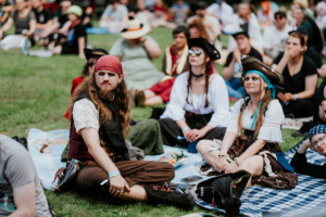 Zuschauer in Piratenkostümen beim Elbenwald Festival, entspannen auf einer Wiese. Die Gruppe genießt die Atmosphäre des Festivals, während sie auf die Bühne schaut. Bunte und kreative Outfits spiegeln die Leidenschaft der Teilnehmer für Fantasy und Musik wider.