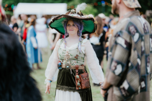 Eine Teilnehmerin des Elbenwald Festivals trägt ein fantasievolles Kostüm mit einem großen Hut und bunten Zöpfen. Im Hintergrund sind weitere Festivalbesucher und festliche Lichter zu sehen, die die lebhafte Atmosphäre des Events unterstreichen.