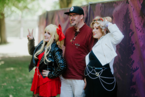 Harmoonics Drei Personen posieren fröhlich vor einem bunten Hintergrund beim Elbenwald Festival. Die Frauen tragen auffällige Cosplay-Outfits, während der Mann in lässiger Kleidung steht. Die Szene spiegelt die kreative und lebendige Atmosphäre des Festivals wider.