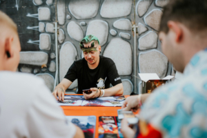 Ein Teilnehmer des Elbenwald Festivals spielt ein Kartenspiel an einem bunten Tisch, während er von anderen Spielern umgeben ist. Im Hintergrund ist eine kreative Wandgestaltung zu sehen, die zur nerdigen Atmosphäre der Veranstaltung beiträgt.
