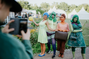 Vier Frauen in bunten Kostümen posieren fröhlich auf dem Elbenwald Festival. Sie tragen kreative Outfits in lebhaften Farben, während im Hintergrund Festivalzelte und Blumen zu sehen sind. Ein Besucher hält eine Kamera, um den Moment festzuhalten.