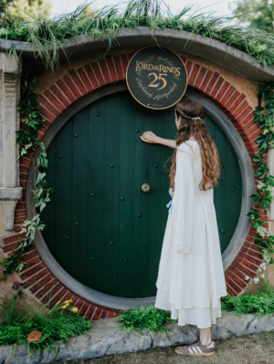 Eine Person in einem langen weißen Kleid klopft an eine grüne Hobbit-Tür, dekoriert mit einem „Der Herr der Ringe“-Schild zum 25. Jubiläum, umgeben von Pflanzen und Blumen, beim Elbenwald Festival, das Musik und Nerd-Kultur vereint.