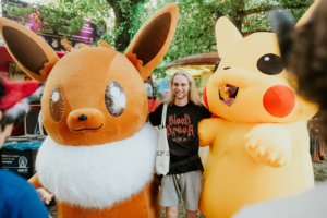 Ein Besucher des Elbenwald Festivals posiert mit übergroßen, plüschigen Charakteren von Pikachu und Evoli. Die lebhaften Farben und die fröhliche Atmosphäre unterstreichen den einzigartigen Mix aus Musik und Nerdkultur auf dem Festival.
