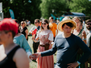 Ein fröhlicher Teilnehmer in historischer Kleidung mit einem Fächer beim Elbenwald Festival, umgeben von einer bunten Menge. Die Szene zeigt die lebendige Atmosphäre des Festivals, das Musik und Nerd-Kultur verbindet.