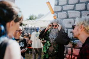 Roleplay Village Ein Teilnehmer des Elbenwald Festivals in einem kreativen Kostüm mit grauem Bart und Sonnenbrille, umgeben von anderen Festivalbesuchern, vor einer fantasievollen Kulisse. Die Atmosphäre ist lebhaft und einladend, perfekt für Fans von Musik und Nerdkultur.