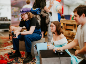 Gamesroom by Nerdistan Zwei Teilnehmer des Elbenwald Festivals spielen Videospiele in Kostümen. Ein junger Mann in blauer Overalls und eine Frau in einem hellblauen Kleid sitzen auf Kisten und genießen die entspannte Atmosphäre des Events, umgeben von anderen Besuchern und Dekoration.