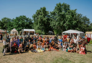 Gruppenfoto von Festivalbesuchern in fantasievollen Kostümen beim Elbenwald Festival. Die Teilnehmer posieren auf einer Wiese vor Zeltständen, umgeben von Bäumen, und zeigen ihre kreativen Outfits, die von verschiedenen Fantasiewelten inspiriert sind.