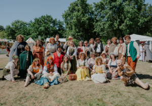 Gruppenszene beim Elbenwald Festival, Teilnehmer in fantasievoller Kleidung posieren auf einer Wiese. Im Hintergrund sind Zelte und Bäume zu sehen, die festliche Atmosphäre unterstreichen. Ideal für Fans von Musik und Fantasie.