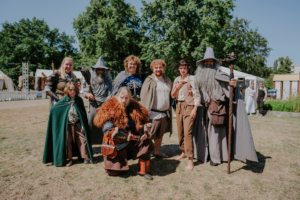 Gruppenszene auf dem Elbenwald Festival mit Teilnehmern in fantasievollen Kostümen, darunter Zauberer, Krieger und Elfen. Die Veranstaltung kombiniert Musik und Nerd-Kultur in einer lebhaften, sommerlichen Festivalatmosphäre.