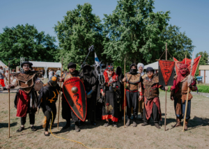 Gruppe von kostümierten Teilnehmern beim Elbenwald Festival, die in beeindruckenden Fantasy-Outfits posieren. Im Hintergrund sind Zelte und Bäume zu sehen, die die festliche Atmosphäre des Festivals unterstreichen.