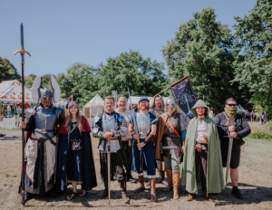 Gruppenszene von Cosplayern beim Elbenwald Festival, gekleidet in fantasievolle Rüstungen und Kostüme. Die Teilnehmer posieren fröhlich im Freien, umgeben von einem bunten Festivalgelände mit Zelten und Bäumen im Hintergrund.