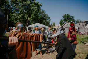 Bunt gekleidete Teilnehmer des Elbenwald Festivals inmitten eines Rollenspiel-Duells. Die Szene zeigt verschiedene kostümierte Charaktere, die mit Waffen in einem Freiluftbereich interagieren, umgeben von einer festlichen Atmosphäre und grüner Natur.