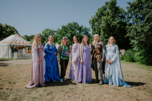 Gruppe von Frauen in fantasievollen Kostümen auf dem Elbenwald Festival, umgeben von festlicher Atmosphäre und Zelt im Hintergrund. Die Teilnehmerinnen verkörpern verschiedene Charaktere und genießen die Mischung aus Musik und Nerdkultur.