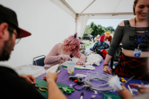 Teilnehmer des Elbenwald Festivals arbeiten an kreativen Cosplay-Projekten an einem Basteltisch. Auf dem Tisch sind Materialien wie Stoff, Scheren und Bastelwerkzeuge sichtbar, während die Atmosphäre von Begeisterung und Kreativität geprägt ist.