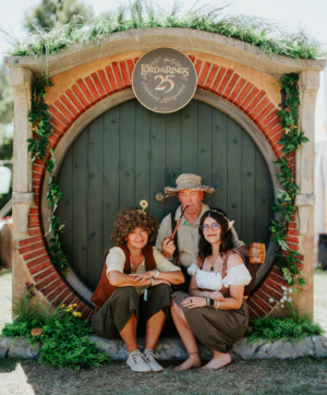 Drei verkleidete Festivalbesucher posieren vor einer Hobbit-Tür mit einem "Der Herr der Ringe"-Schild. Die Szene spiegelt die kreative Atmosphäre des Elbenwald Festivals wider, wo Musik und Fantasie aufeinandertreffen.