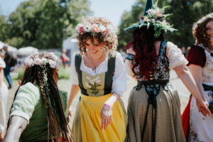 Bunt gekleidete Festivalbesucherinnen beim Elbenwald Festival tanzen und lachen im Freien. Die Frauen tragen Blumenkränze und traditionelle Gewänder, während sie die sommerliche Atmosphäre und die Gemeinschaft genießen.