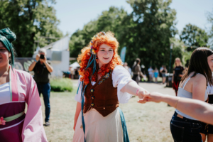 Zwei Frauen in Kostümen interagieren fröhlich auf dem Elbenwald Festival. Die Frau mit lockigem, rotem Haar trägt ein historisches Outfit und reicht einer anderen Person die Hand. Im Hintergrund sind weitere Festivalbesucher und Stände zu sehen.